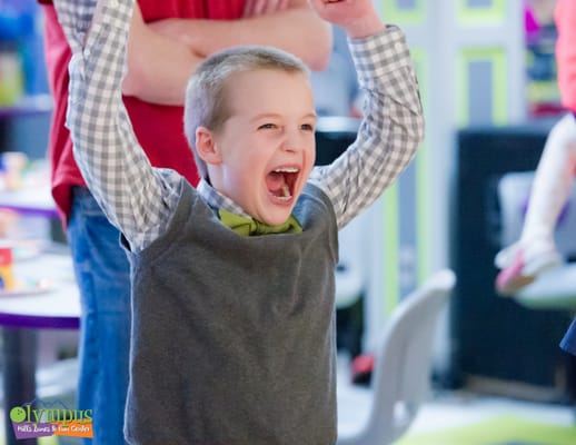 A young boy in a grey vest and bowtie cheers with his arms raised at a bowling center.