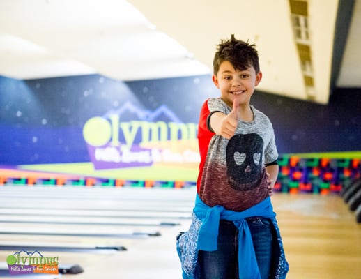 A smiling young boy giving a thumbs-up in front of bowling lanes.