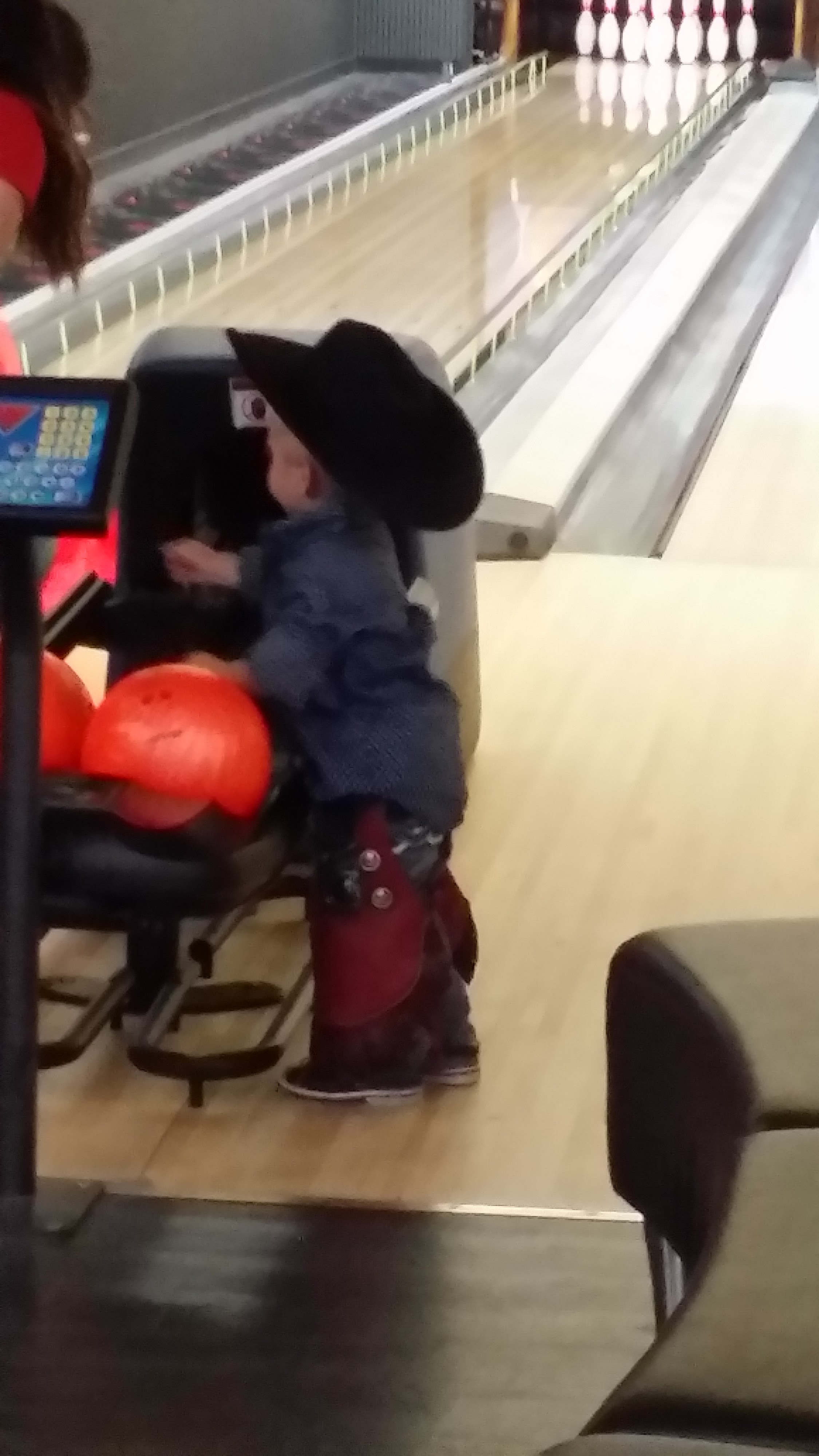 A young child in a cowboy hat stands next to orange bowling balls at a bowling lane with bumpers.