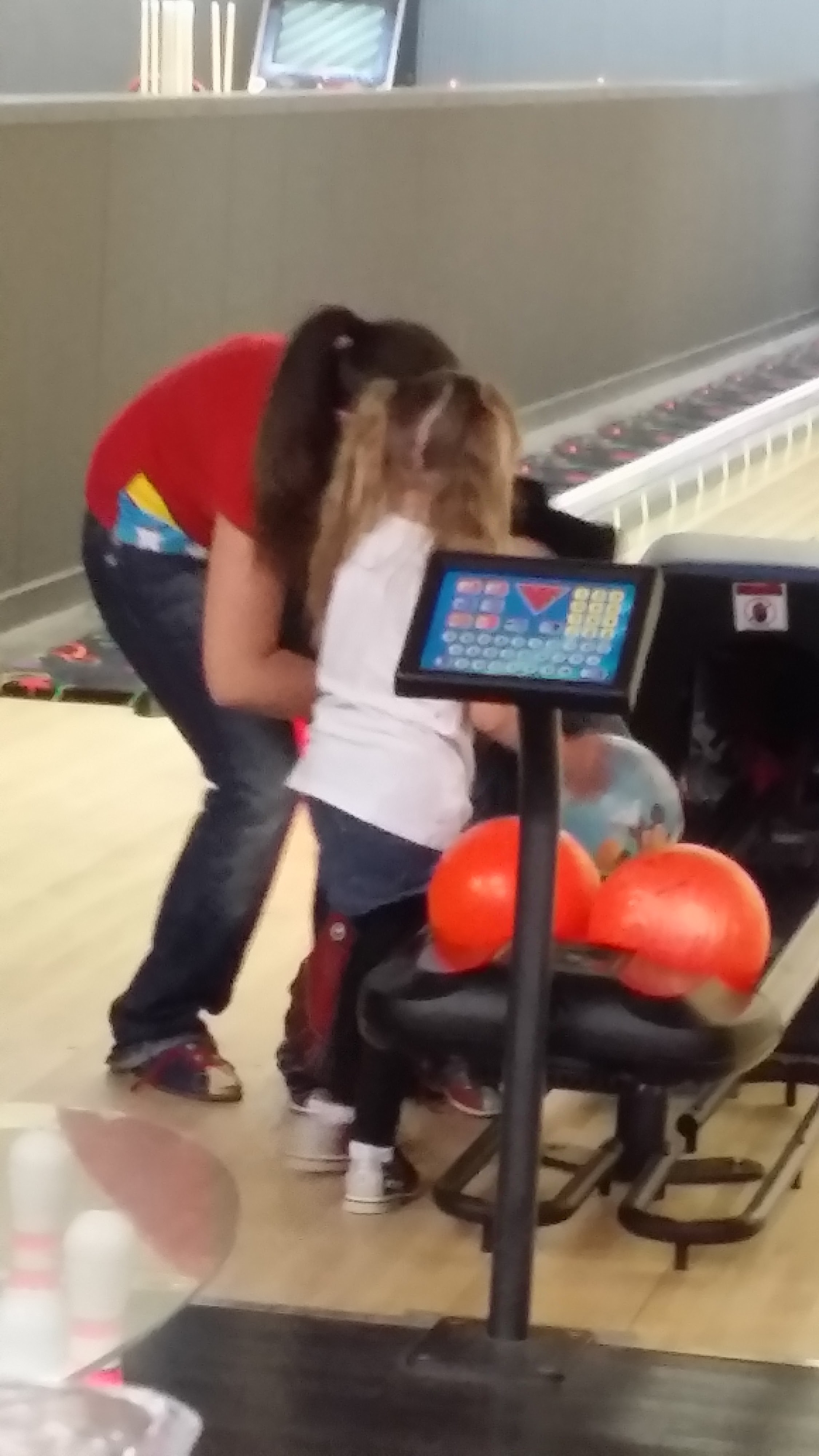 A woman in a red shirt helps a small child with a bowling ball next to a scoring tablet at the lanes.