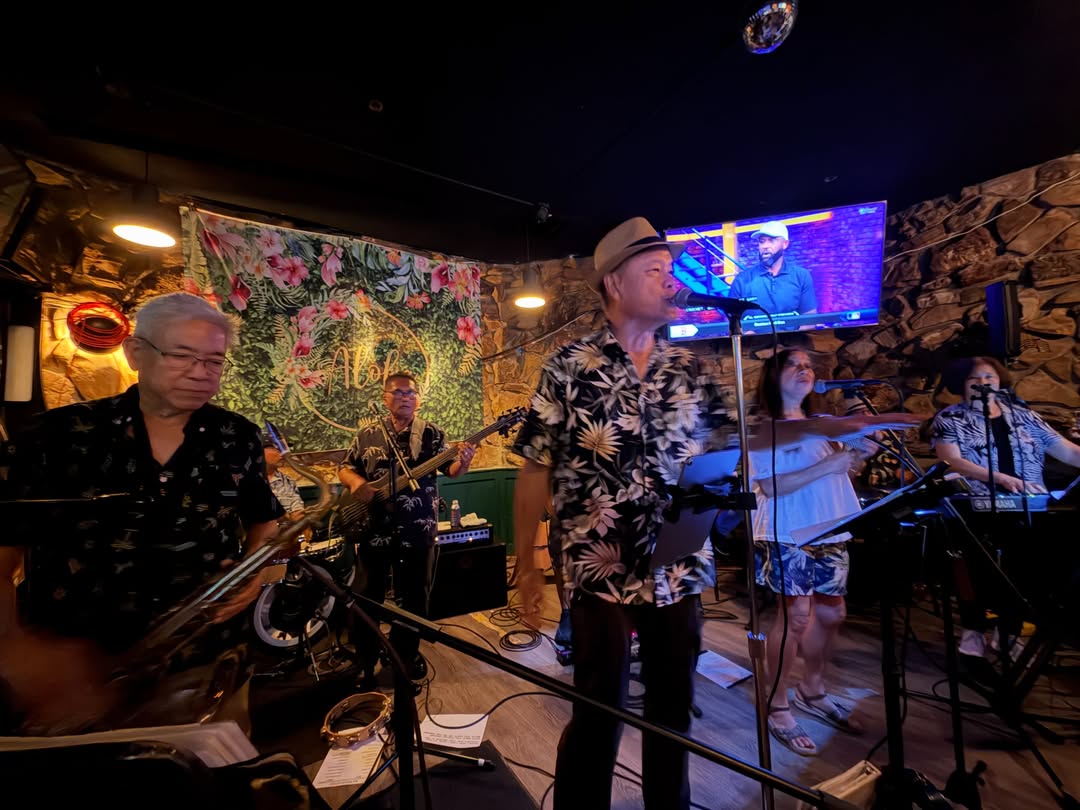 A live band with a singer in a floral shirt performing on a stage in a lounge with stone walls.