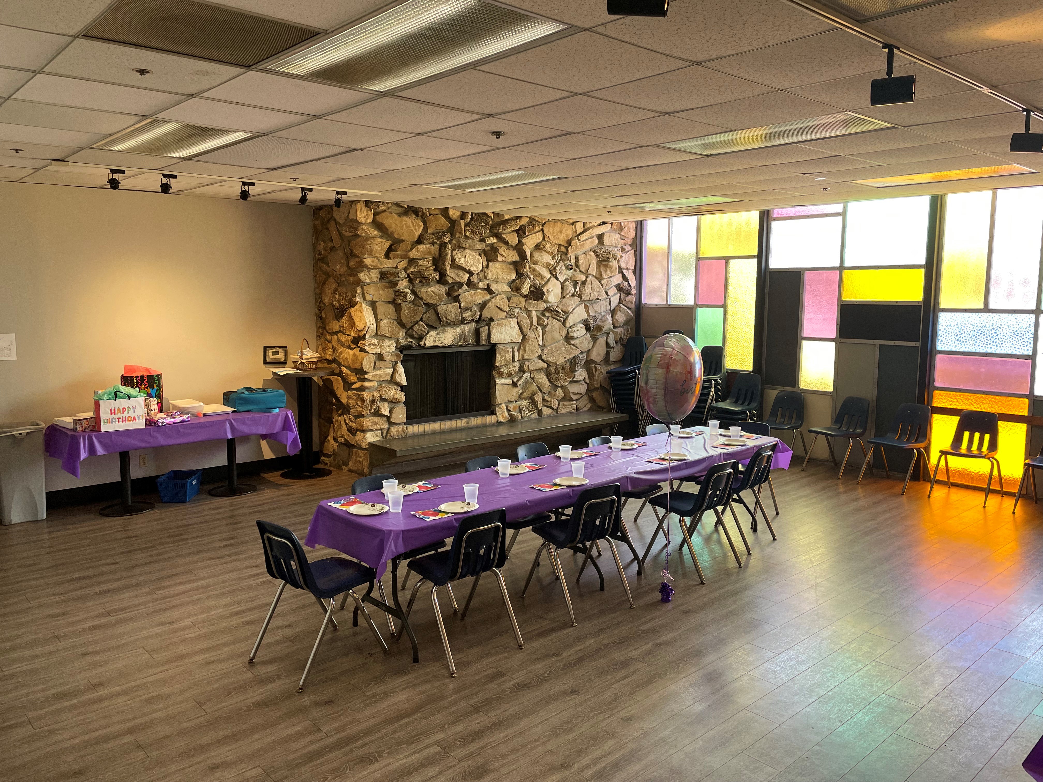 A private party room with a purple-clothed long table, a large stone fireplace, and colorful window panels.