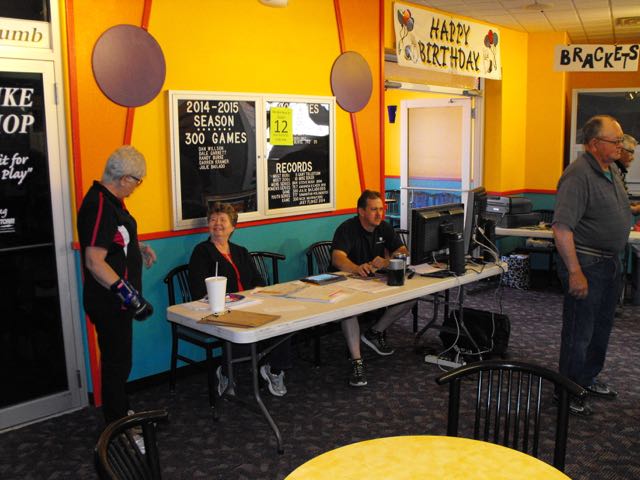 People gather at a league registration desk inside a bowling center with colorful yellow and orange walls.