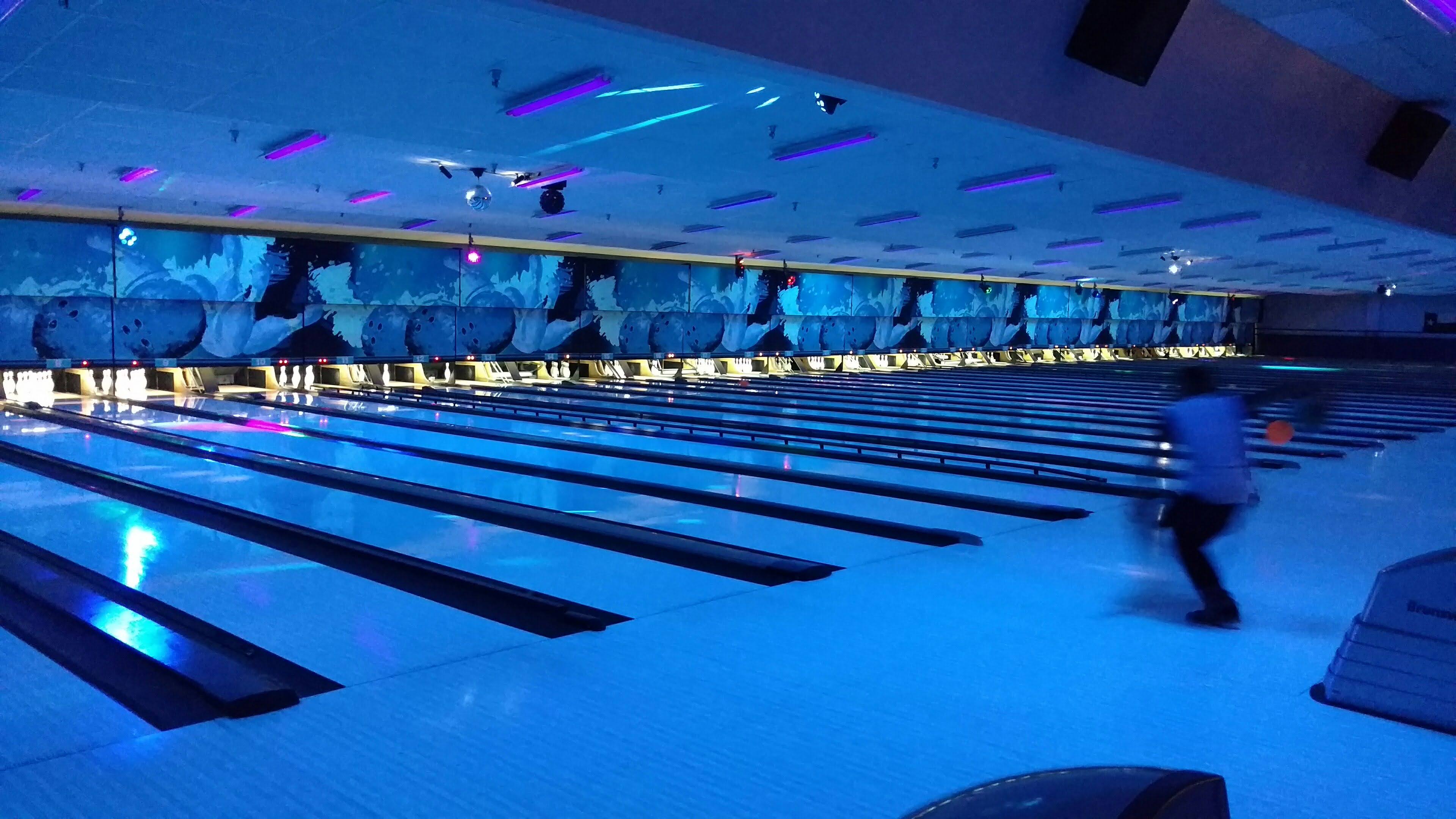 A wide view of several bowling lanes glowing blue under cosmic blacklights with a blurred person bowling.