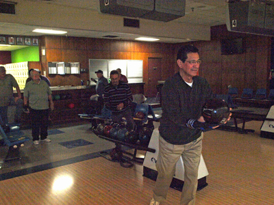 A man in a dark sweater and khakis prepares to bowl at a wood-paneled bowling alley with other men standing nearby.