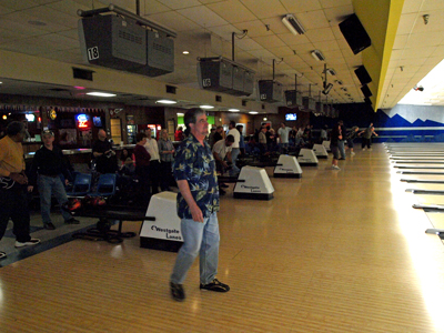 A man walks across a busy bowling alley approach while other players wait by their respective lanes.