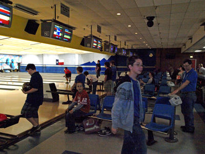 A group of teenagers and children bowling in an indoor center with blue chairs and overhead score monitors.