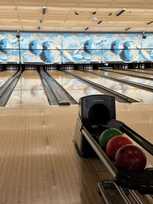 A vertical view of bowling lanes with a ball return holding three colored bowling balls in the foreground.