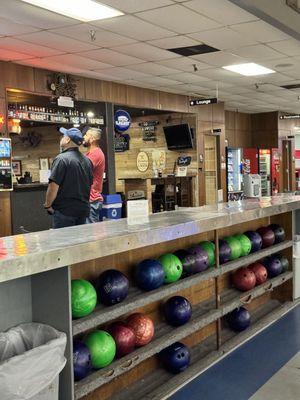 A rack of colorful house bowling balls sits in front of a wood-paneled bar and lounge area.