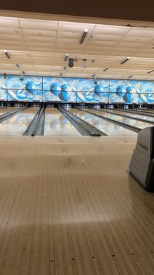 A vertical view looking down polished wooden bowling lanes toward blue decorative back walls and pin setters.