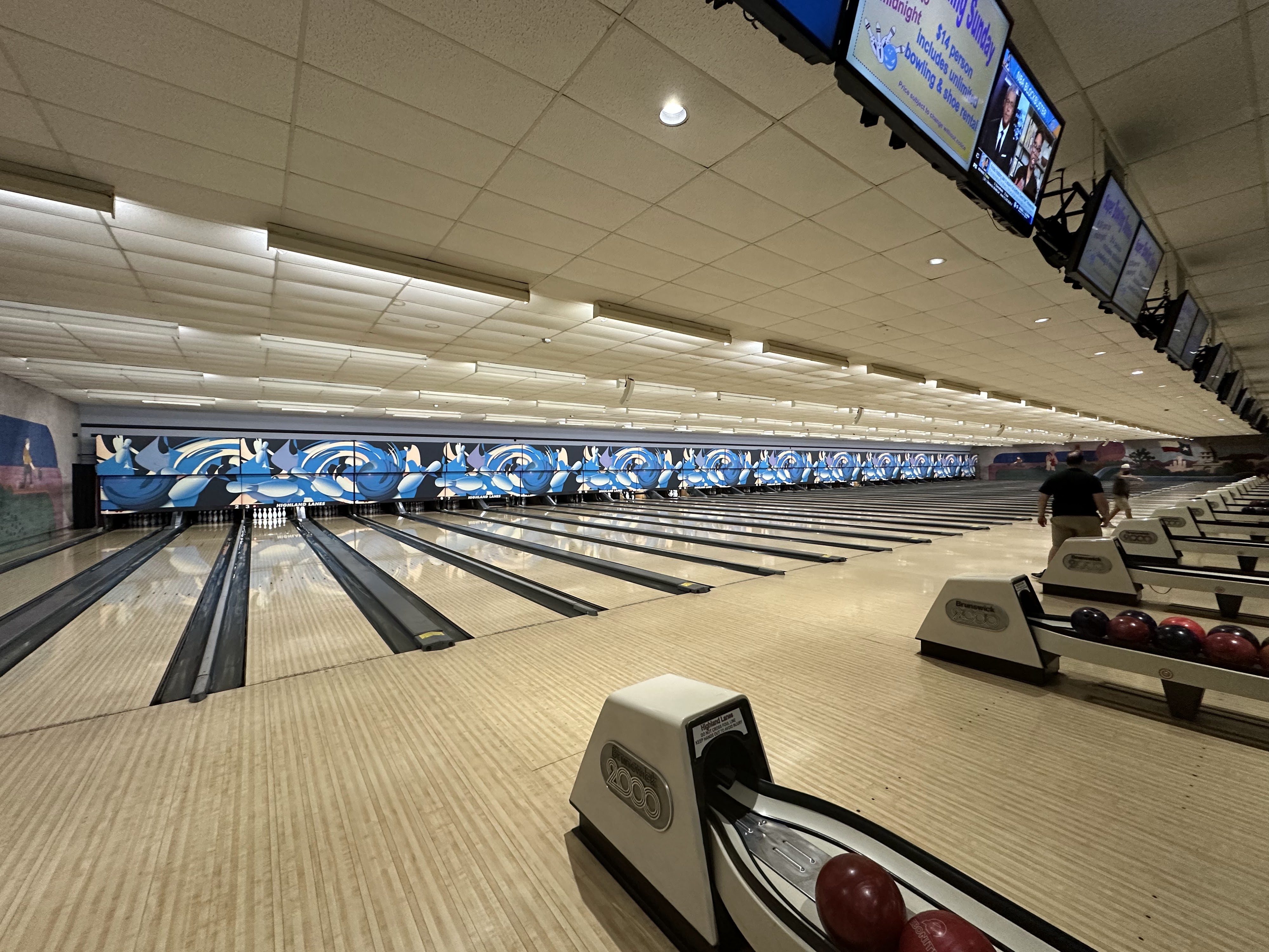 Wide view of numerous bowling lanes with blue wave graphics and overhead monitors at Highland Lanes in Austin.
