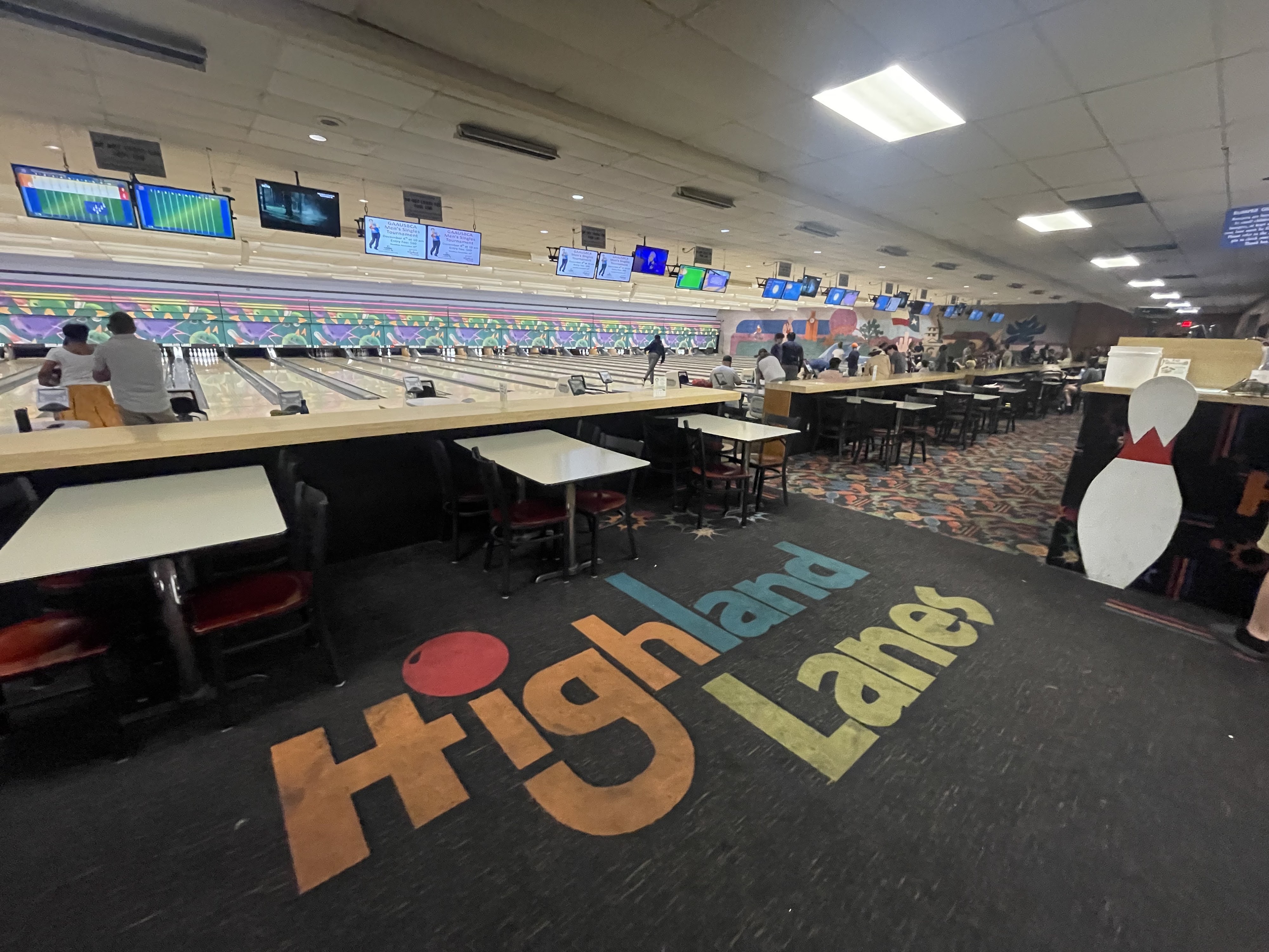 Interior view of a bowling alley with a large colorful logo on the carpet and people playing on the lanes.