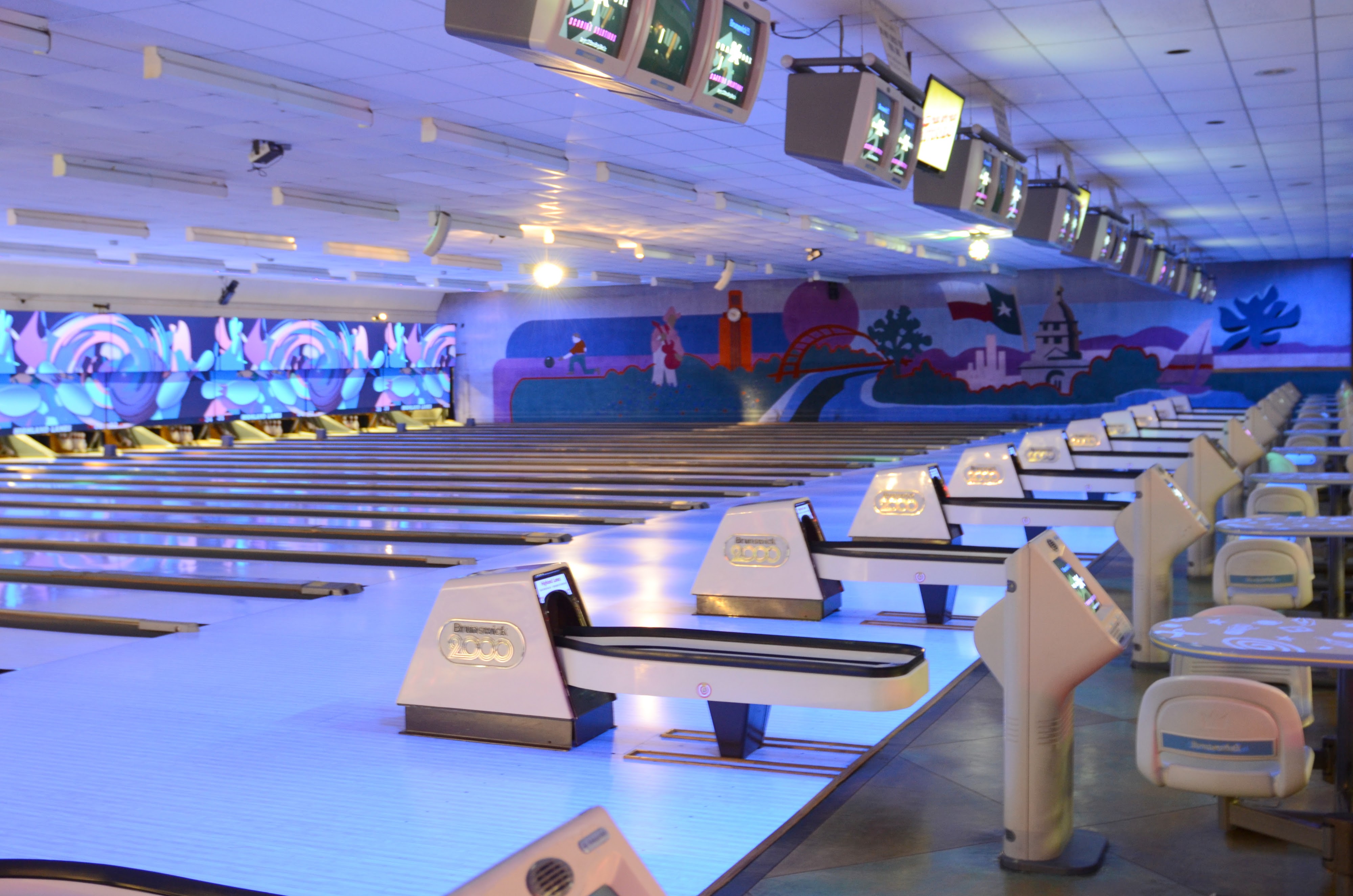 A wide view of many bowling lanes under blue glow lighting with an Austin-themed mural on the back wall.