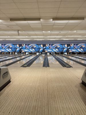 A vertical view of multiple polished bowling lanes leading to pins with blue wave graphics on the back wall.