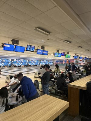 A crowded bowling alley concourse with people socializing at tables and active lanes in the background.