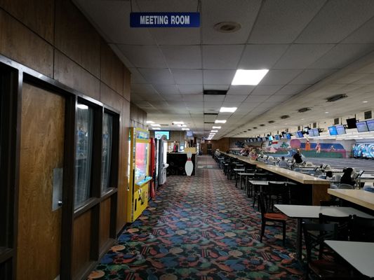 Interior concourse of a bowling alley featuring a meeting room sign, dining tables, and lanes in the background.