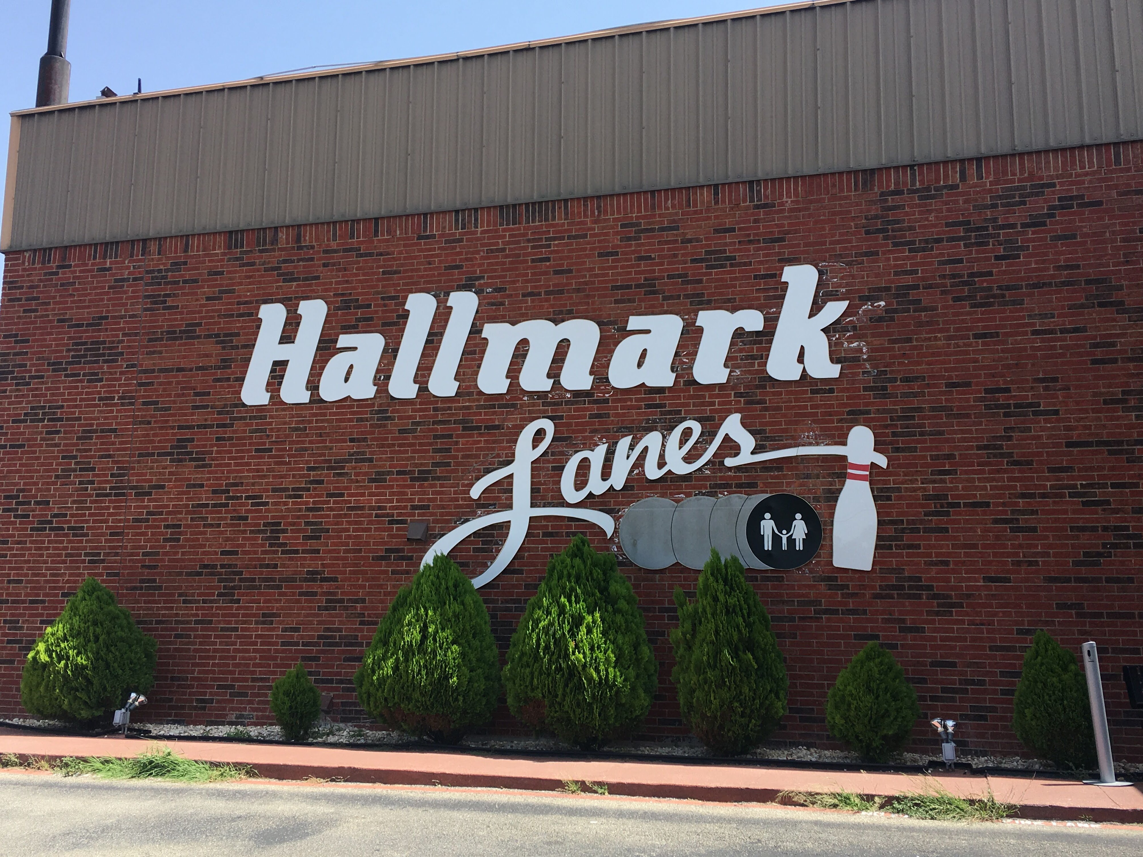 White Hallmark Lanes signage mounted on a red brick exterior wall with small green shrubs and a blue sky.