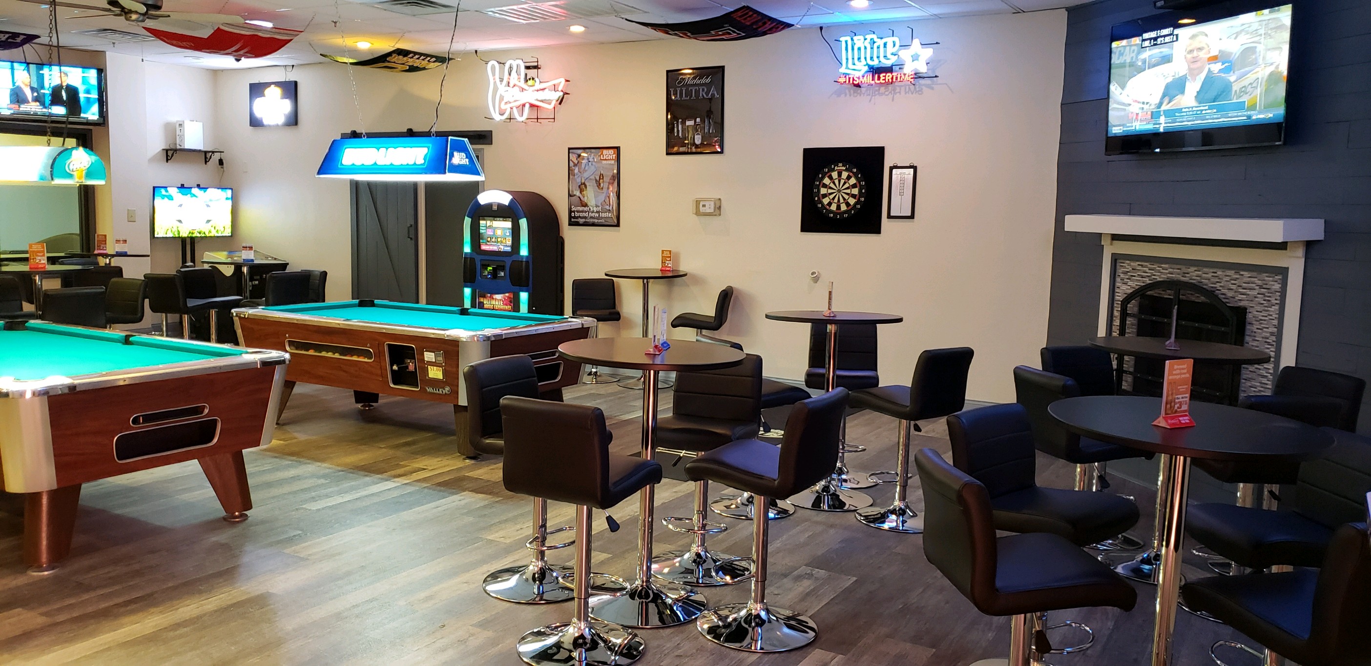 A lounge area with two billiards tables, high-top seating, a jukebox, and televisions mounted on the walls.