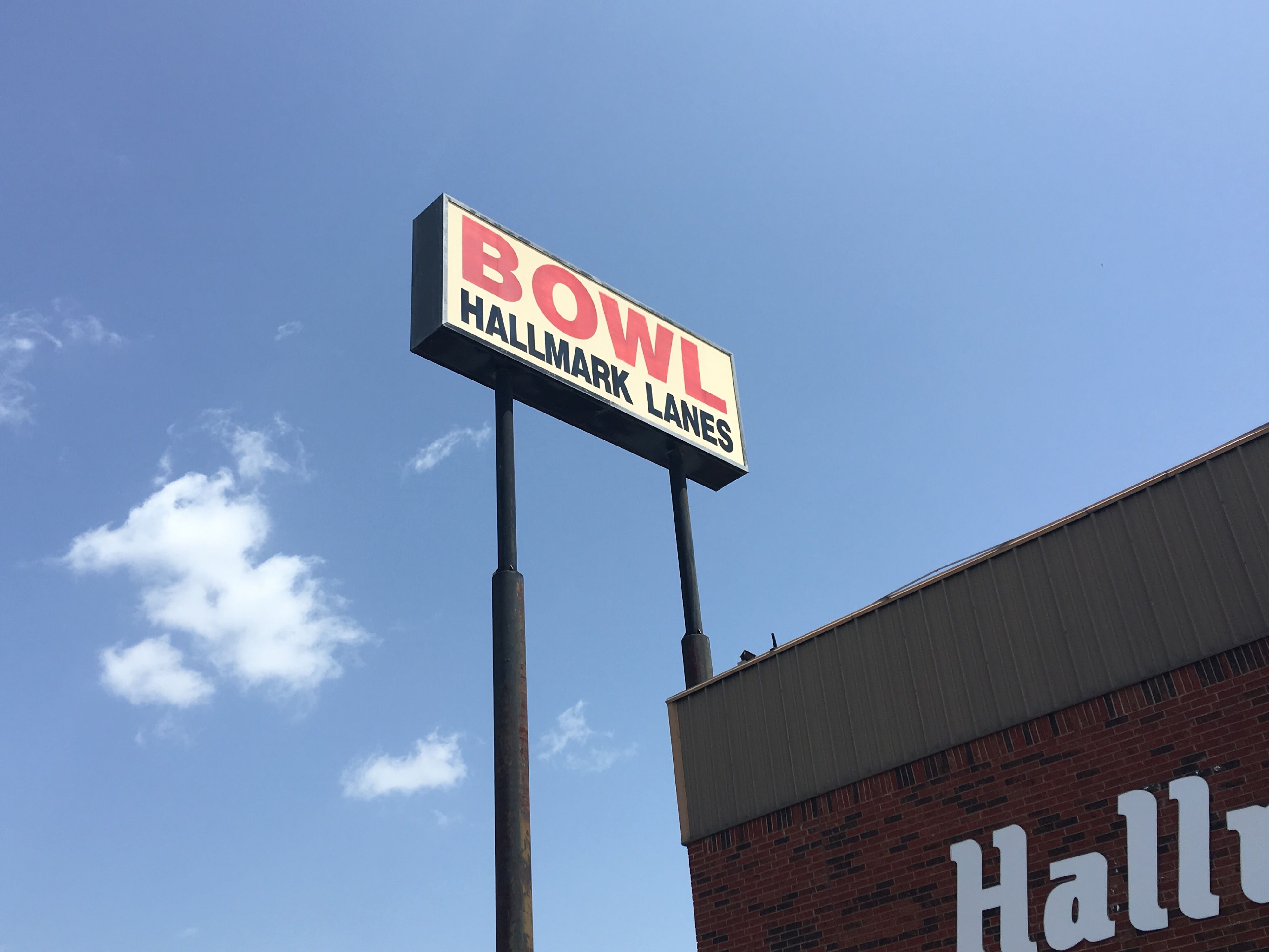 A roadside sign for Hallmark Lanes with the word BOWL in red letters against a bright blue sky.