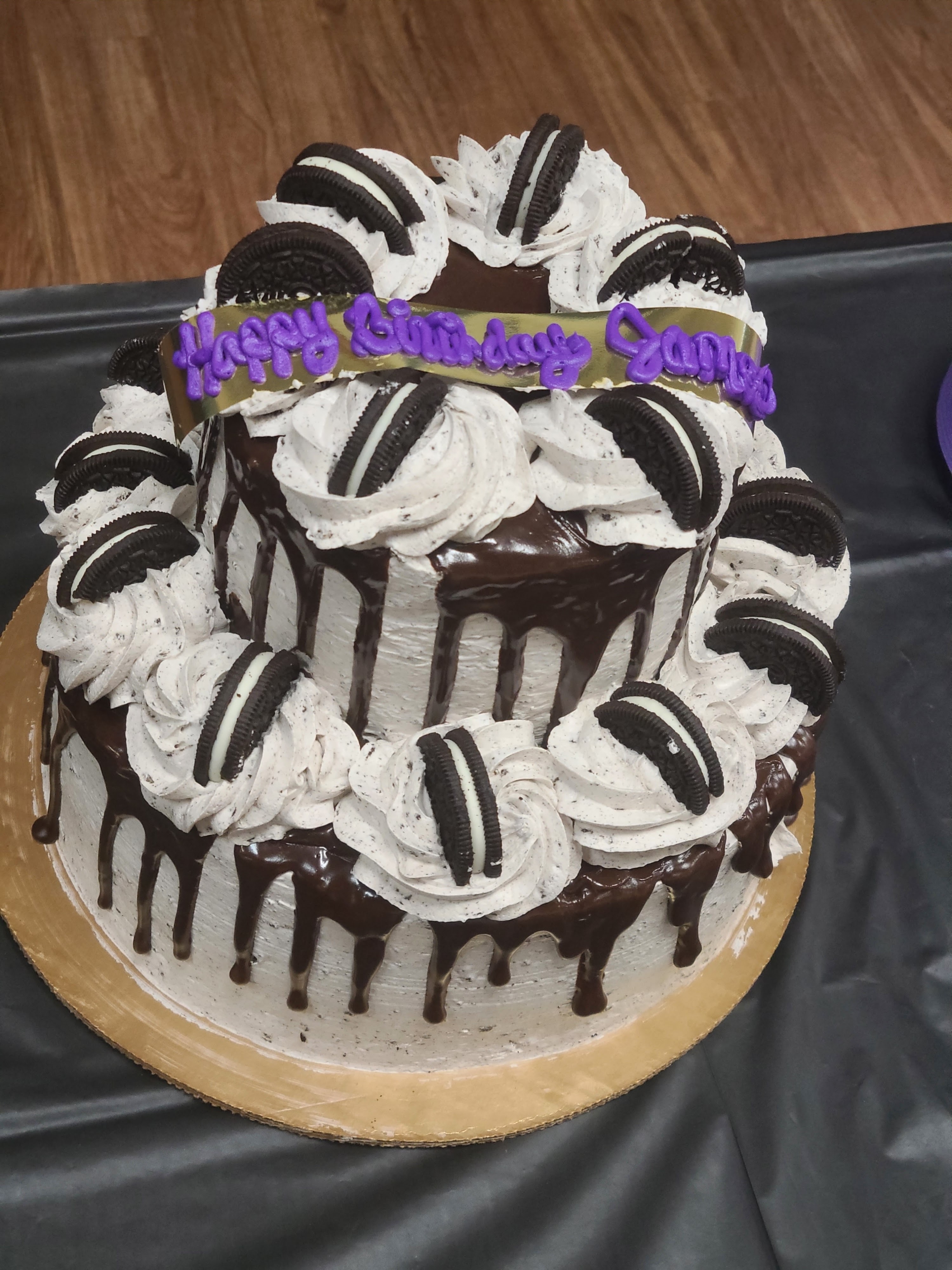 A two-tiered cookies and cream birthday cake with chocolate drip and sandwich cookies on a black table.