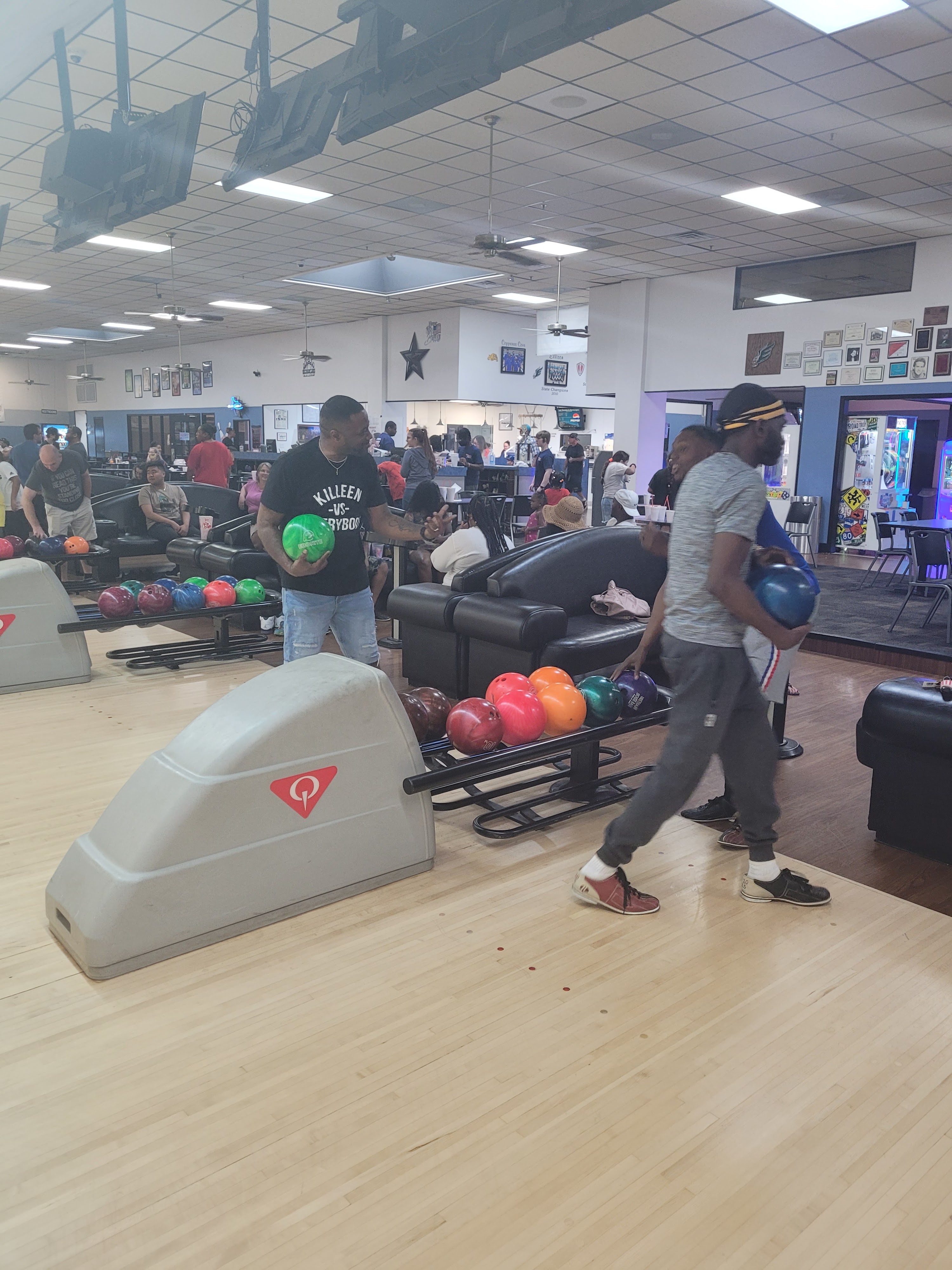 Bowlers holding colorful bowling balls on the lanes near black leather seating areas.