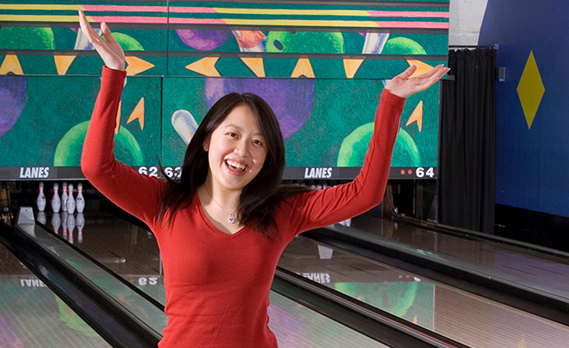 A woman in a red shirt celebrates with her arms raised in front of bowling lanes.