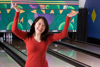 A woman in a red shirt celebrates with her arms raised in front of bowling lanes.