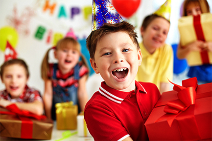 A happy boy wearing a party hat holds a large red gift box while celebrating with friends at a birthday party.