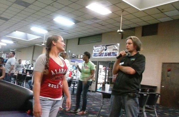 A man speaks into a microphone while standing next to a woman in a red shirt in a bowling center lounge.