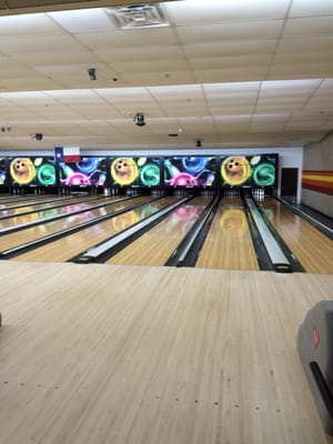 View of multiple polished bowling lanes with overhead scoring monitors and a Texas flag in the background.
