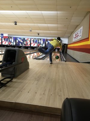 A man in a yellow shirt releases a bowling ball down a wooden lane in a well-lit bowling center.
