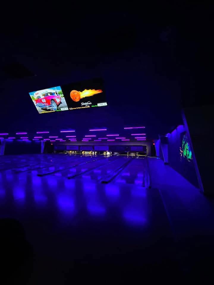 A bowling alley at night, with purple glow lighting illuminating the lanes and screens showing bowling animations.