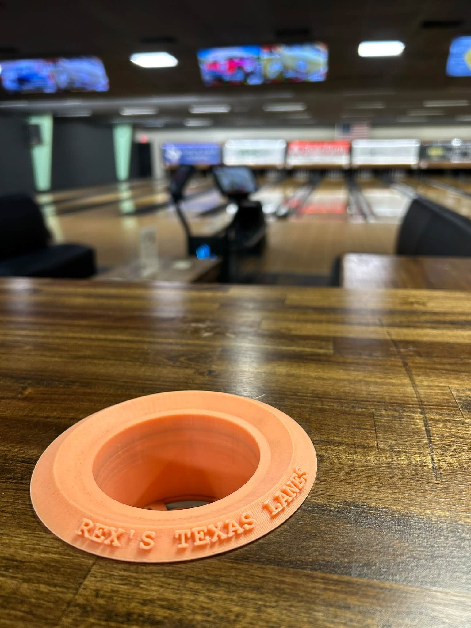 A close-up of a wooden bowling lane table with an orange drink holder featuring "Rex's Texas Lanes" text, with blurred bowling lanes and scoring monitors in the background.
