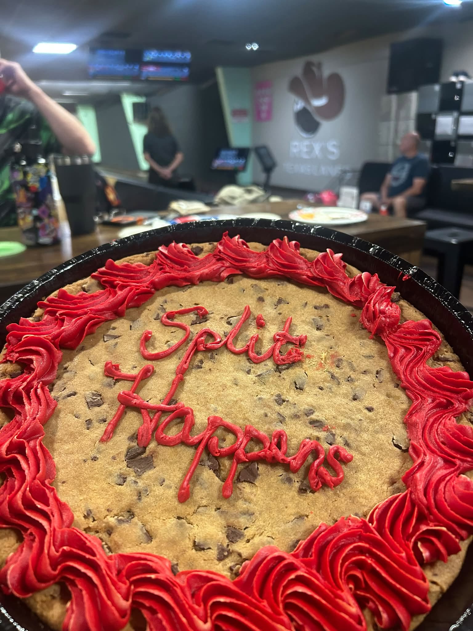 A close-up of a chocolate chip cookie cake with red icing spelling "Split Happens" in a bowling alley background.