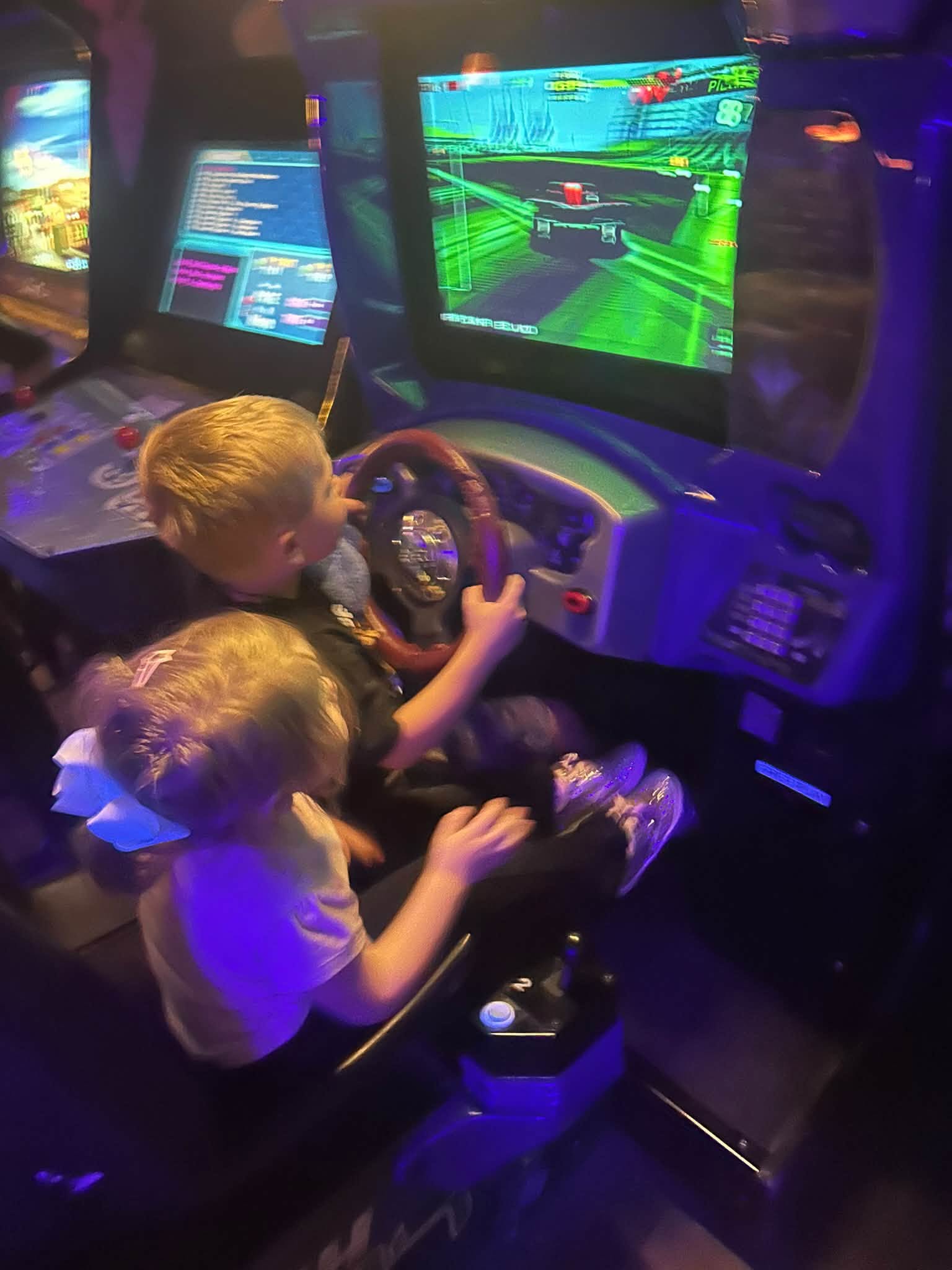 Two children are engaged in a racing arcade game, illuminated by the bright screens in a dimly lit room.