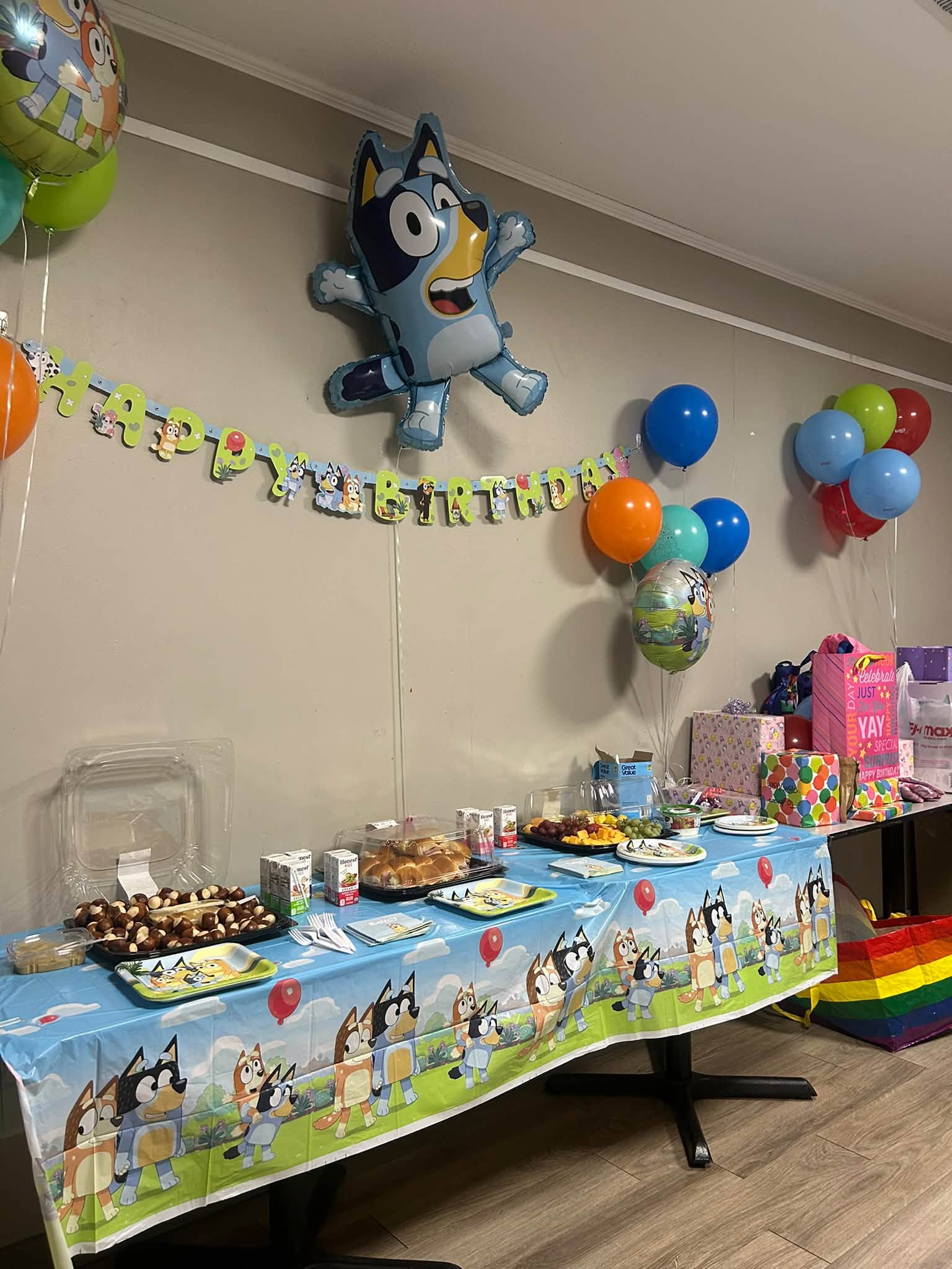 A table set for a "Bluey" themed birthday party, adorned with balloons, a banner, food, and gifts.
