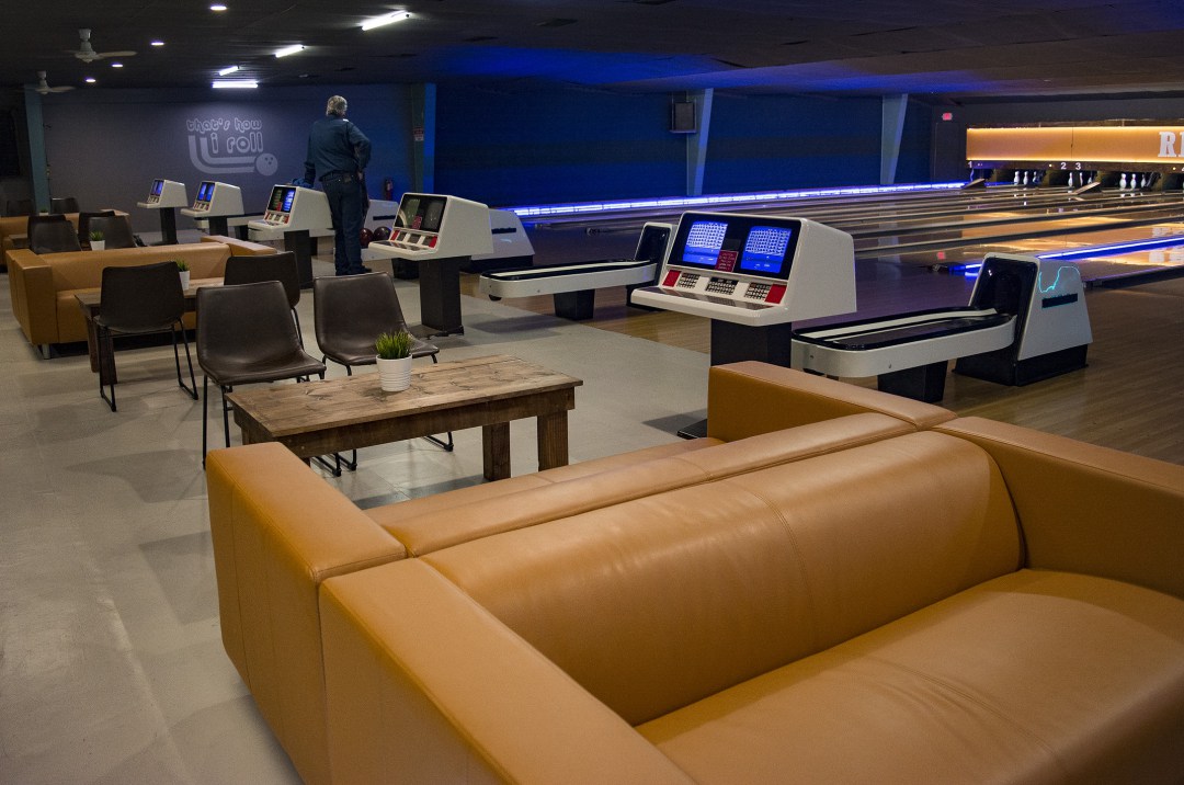 An interior view of a bowling alley with tan couches, dark chairs, and multiple bowling lanes under blue accent lighting.