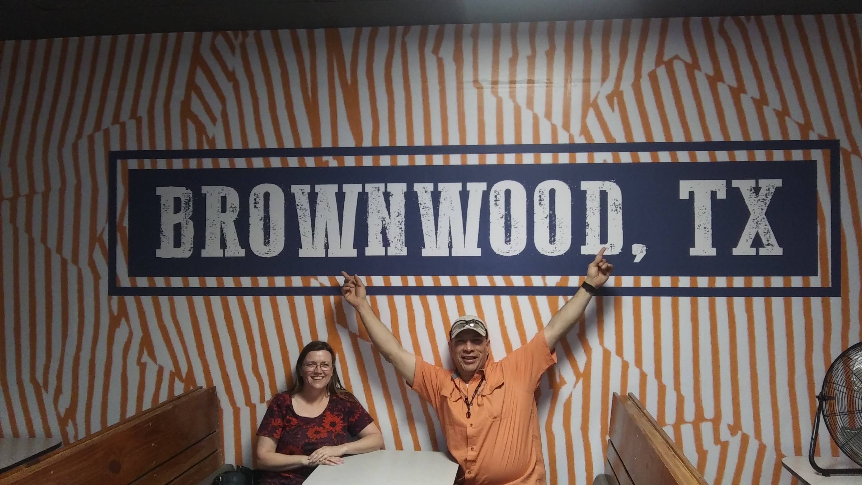 A man and a woman smile at tables in front of a striped wall mural displaying "BROWNWOOD, TX".