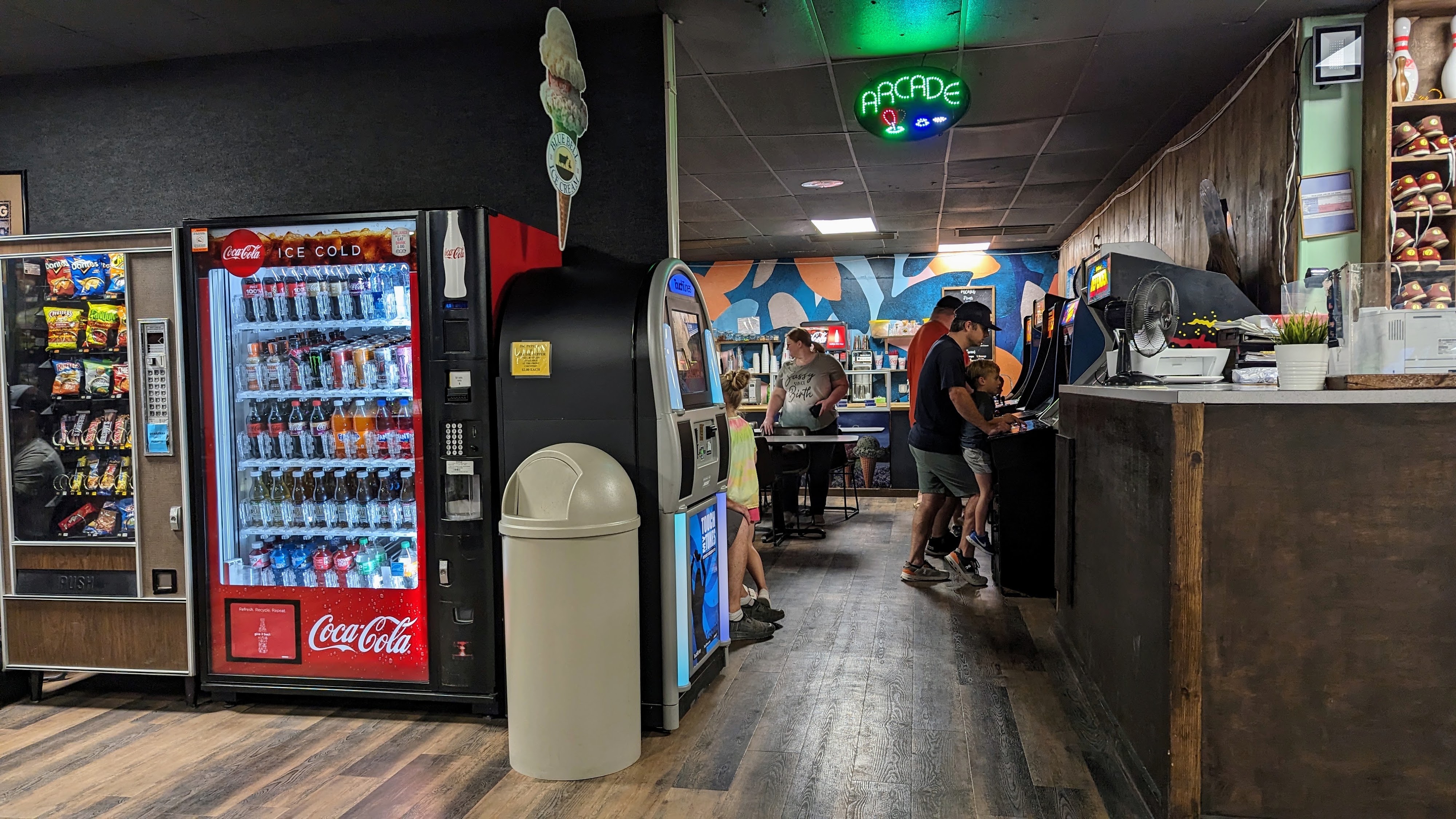 An indoor bowling alley hallway with vending machines, an arcade area with people playing games, and a shoe rental counter.