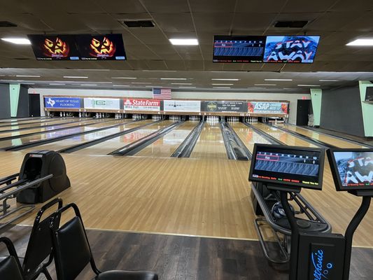 A wide view of multiple bowling lanes with pins set up and overhead scoring displays showing game information and advertisements.
