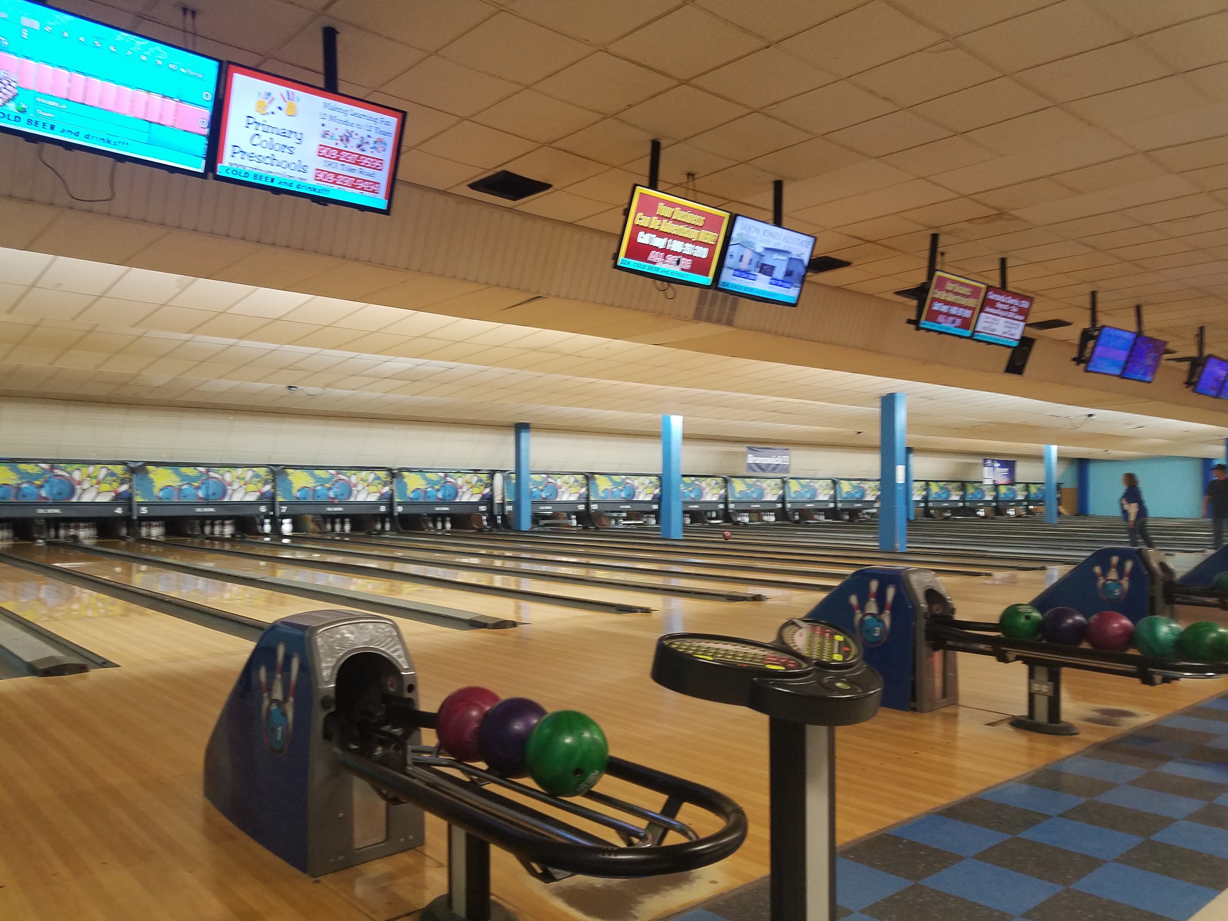 A wide view of several bowling lanes with colorful bowling balls on returns and overhead scoring monitors.