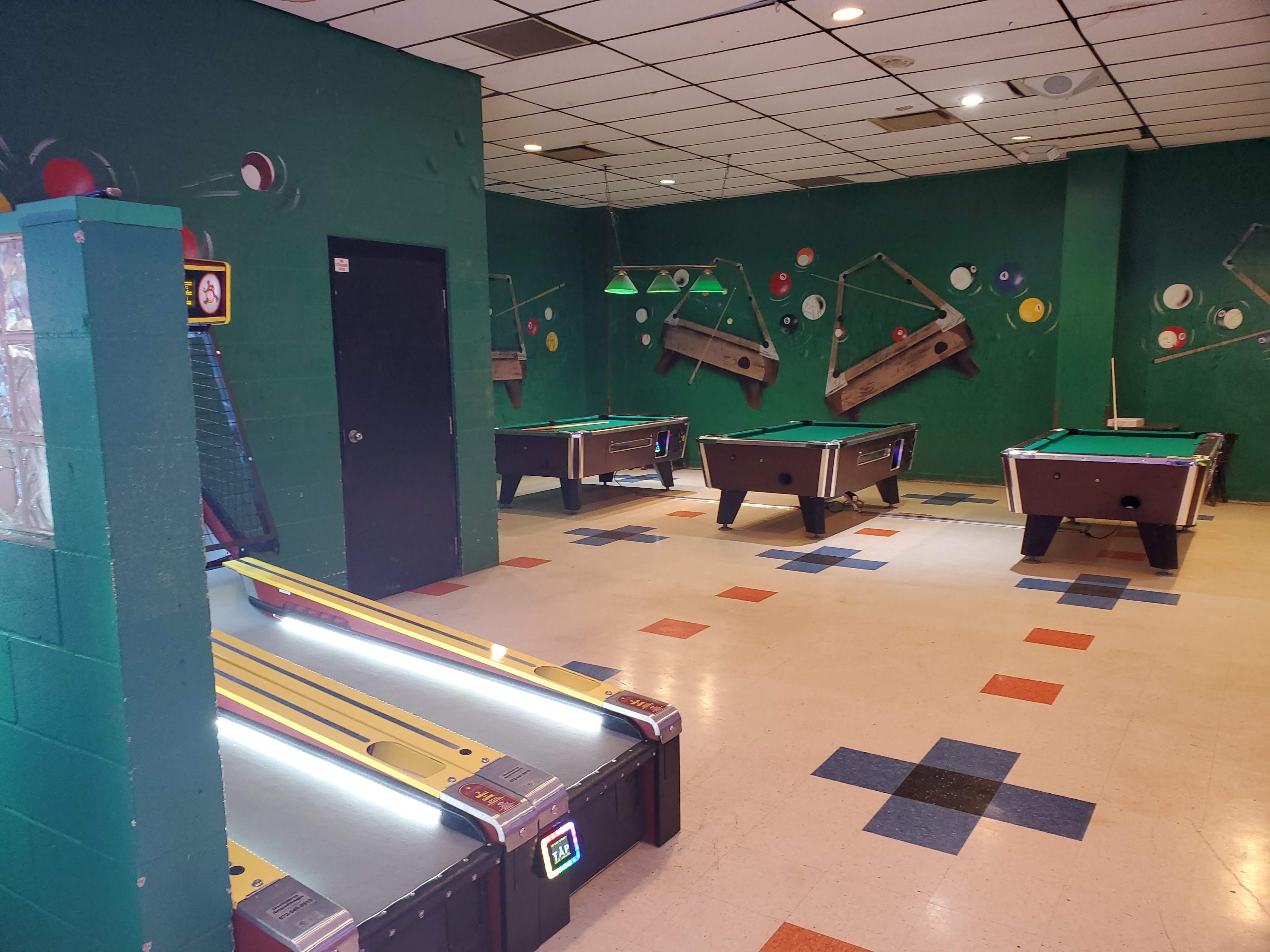A game room with three green pool tables and a skee-ball machine against green walls with pool-themed murals.