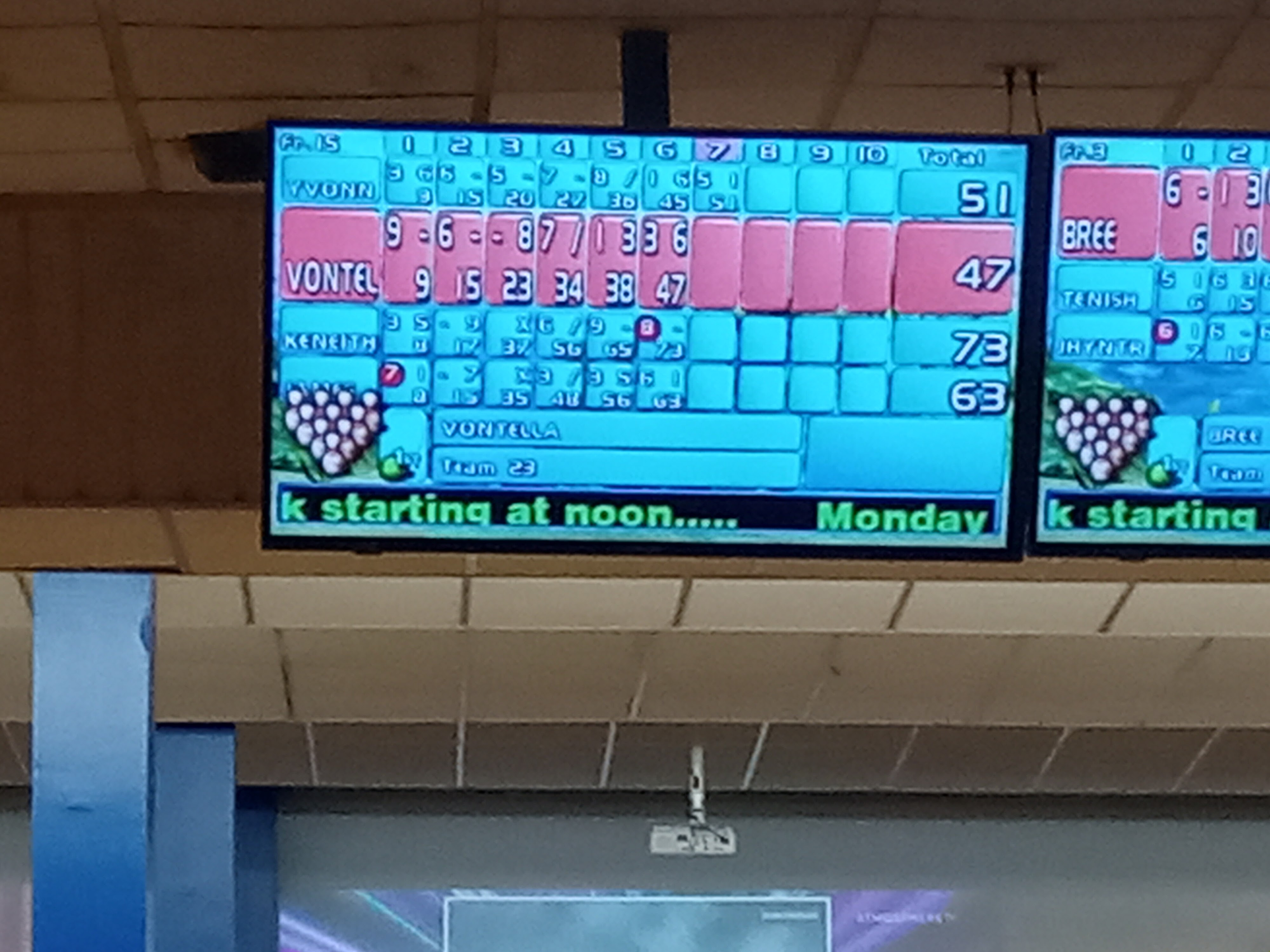 A digital bowling score monitor displaying player names and frame scores hanging from the ceiling.