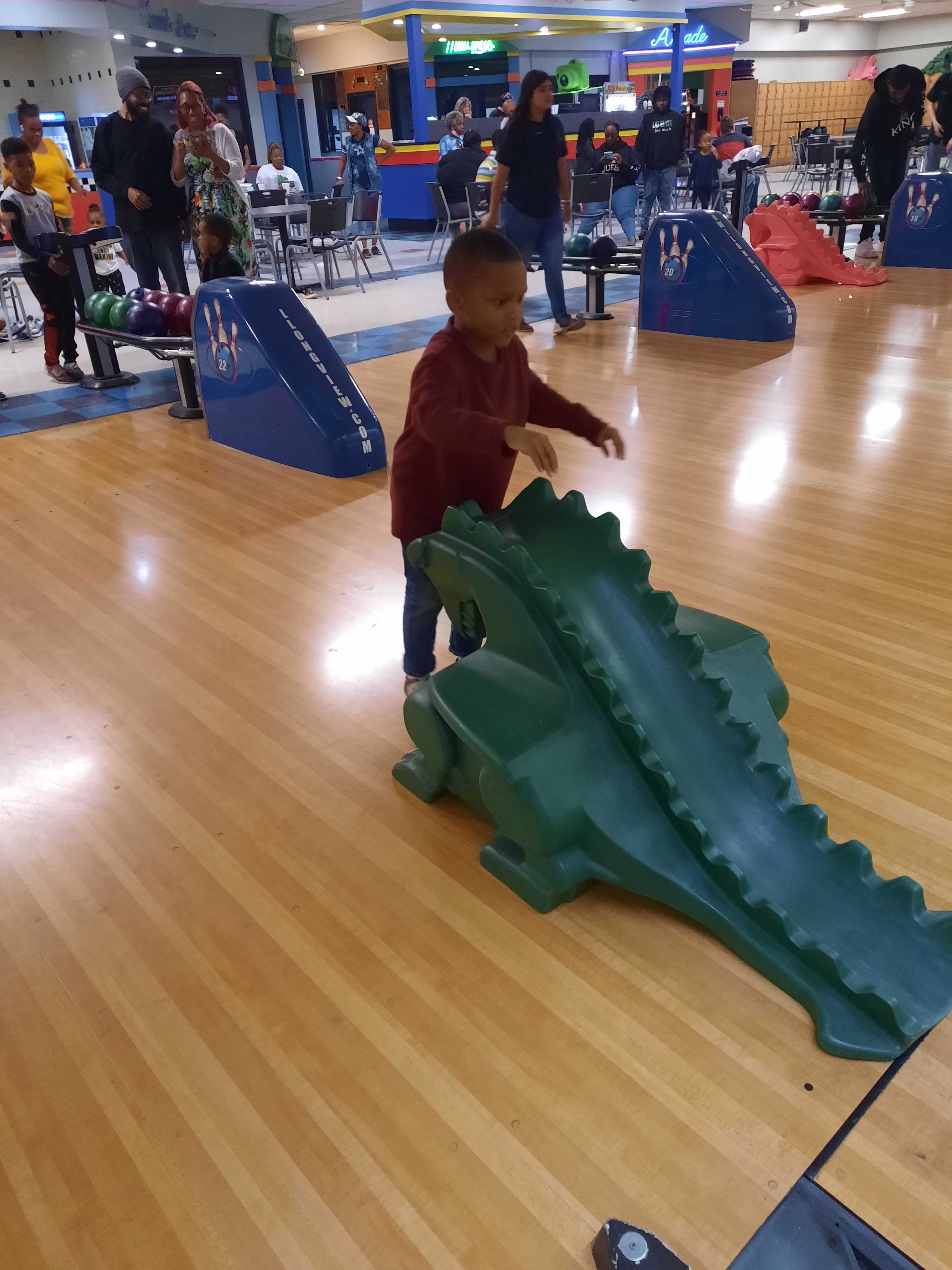 A young boy prepares to use a green dragon-shaped bowling ramp on a lane at a busy bowling center.