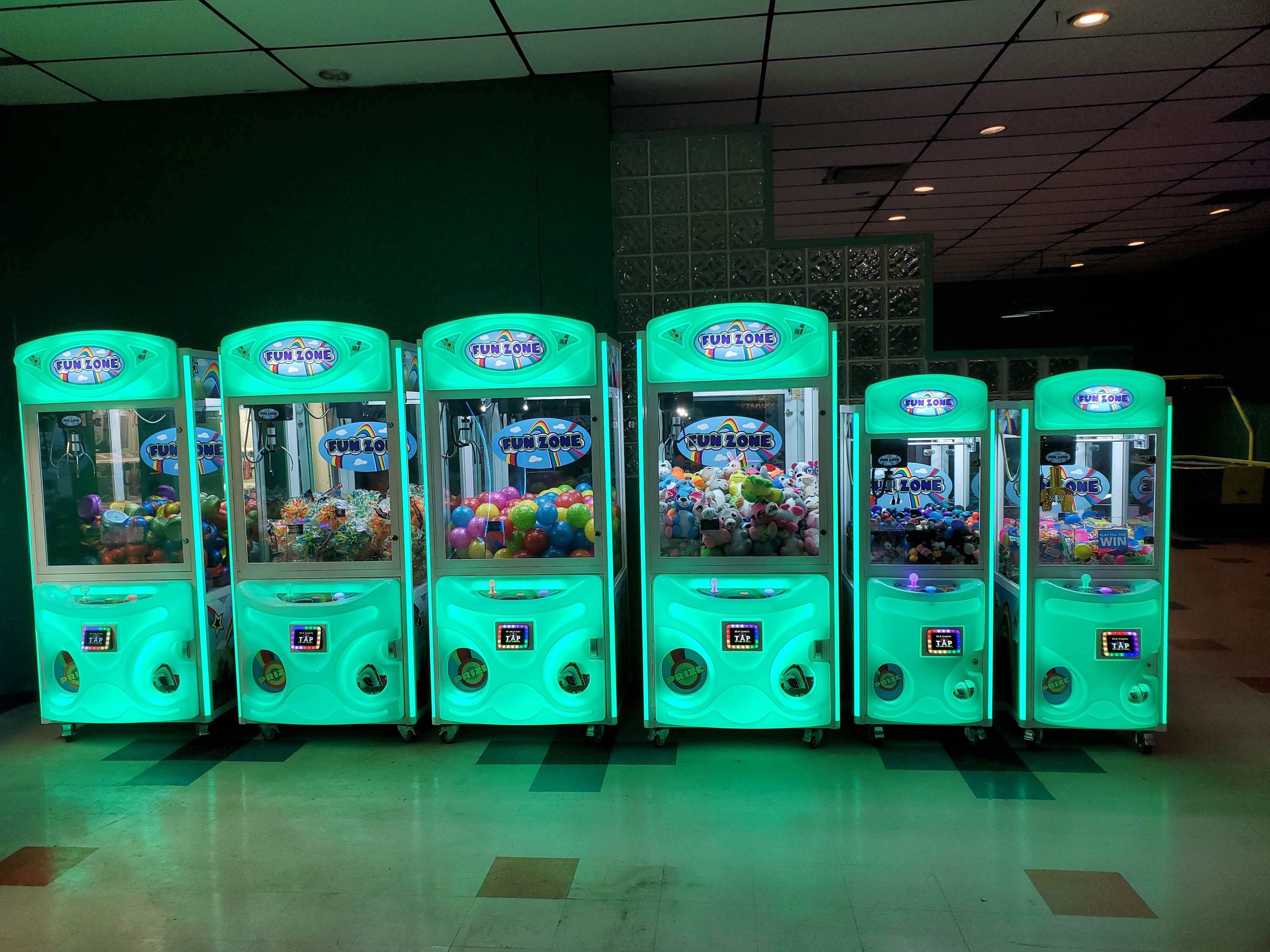 Six brightly lit teal claw machines filled with prizes stand in a row against a dark wall.