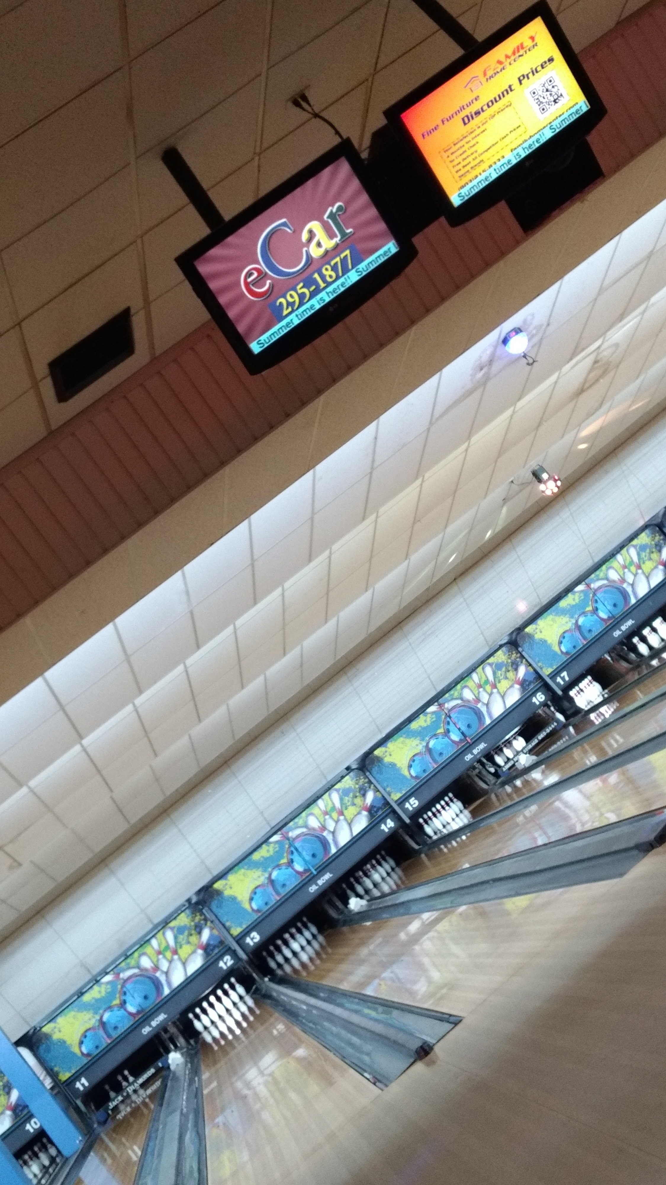 A diagonal view of several bowling lanes with pins set up and digital monitors displaying ads overhead.