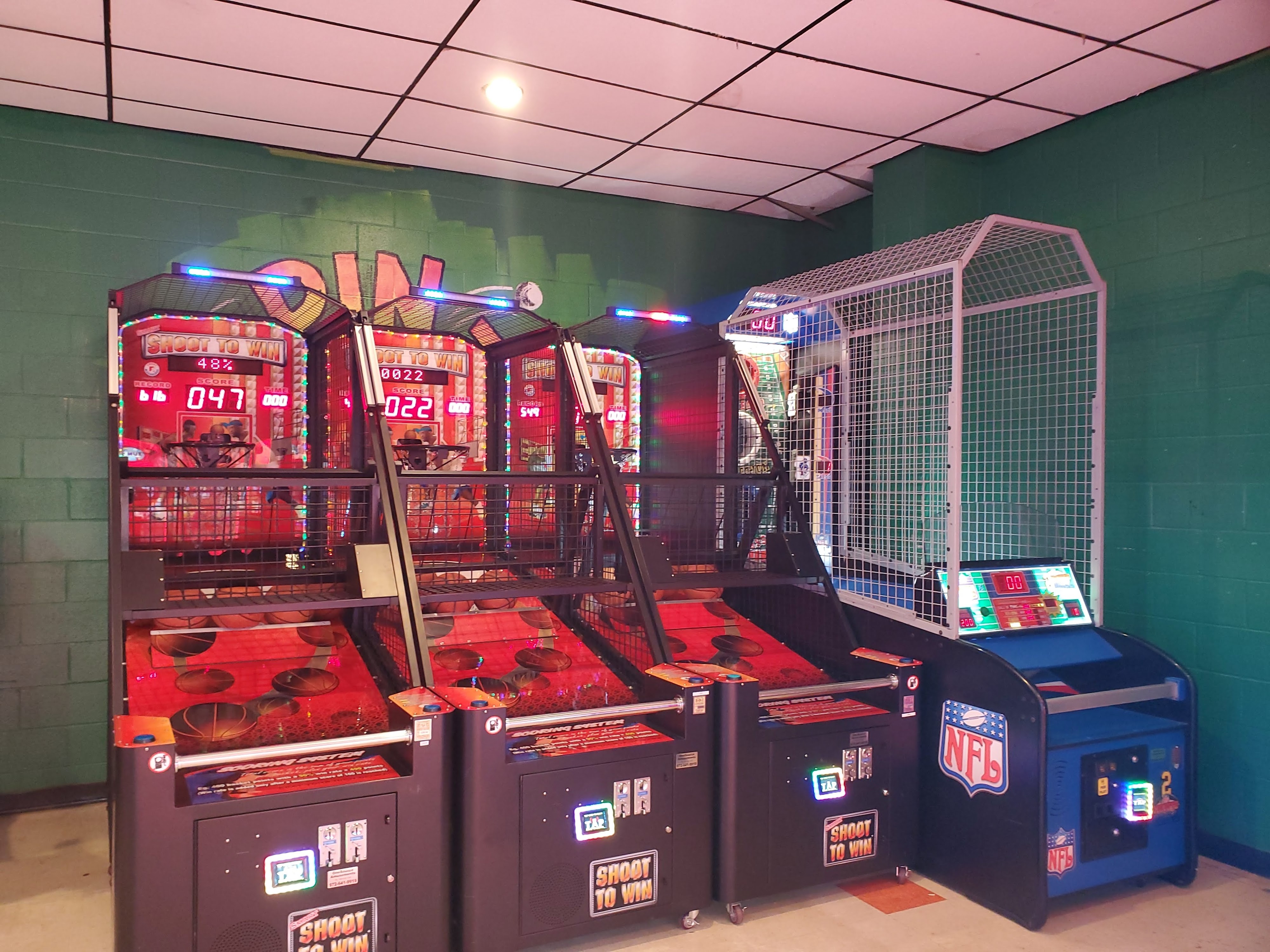 A row of three basketball arcade games and one NFL football toss machine against a green wall.