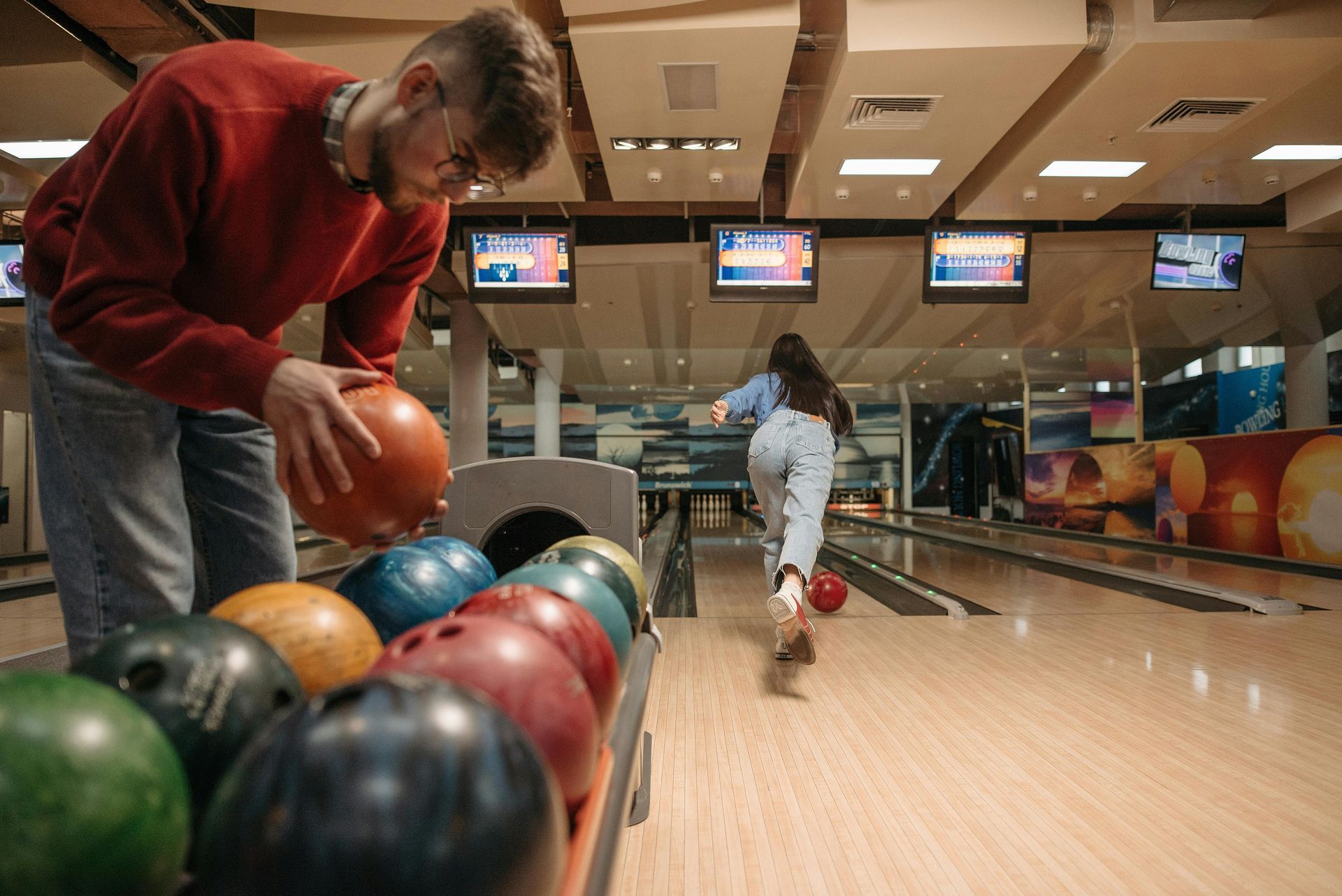 A man picks up a bowling ball while a woman rolls a ball down the lane at a bowling center.
