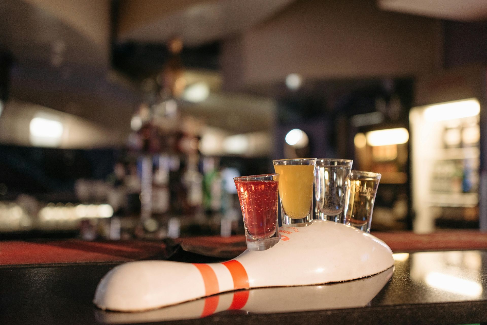 Four shot glasses with different colored liquids in a novelty bowling pin-shaped holder on a bar.