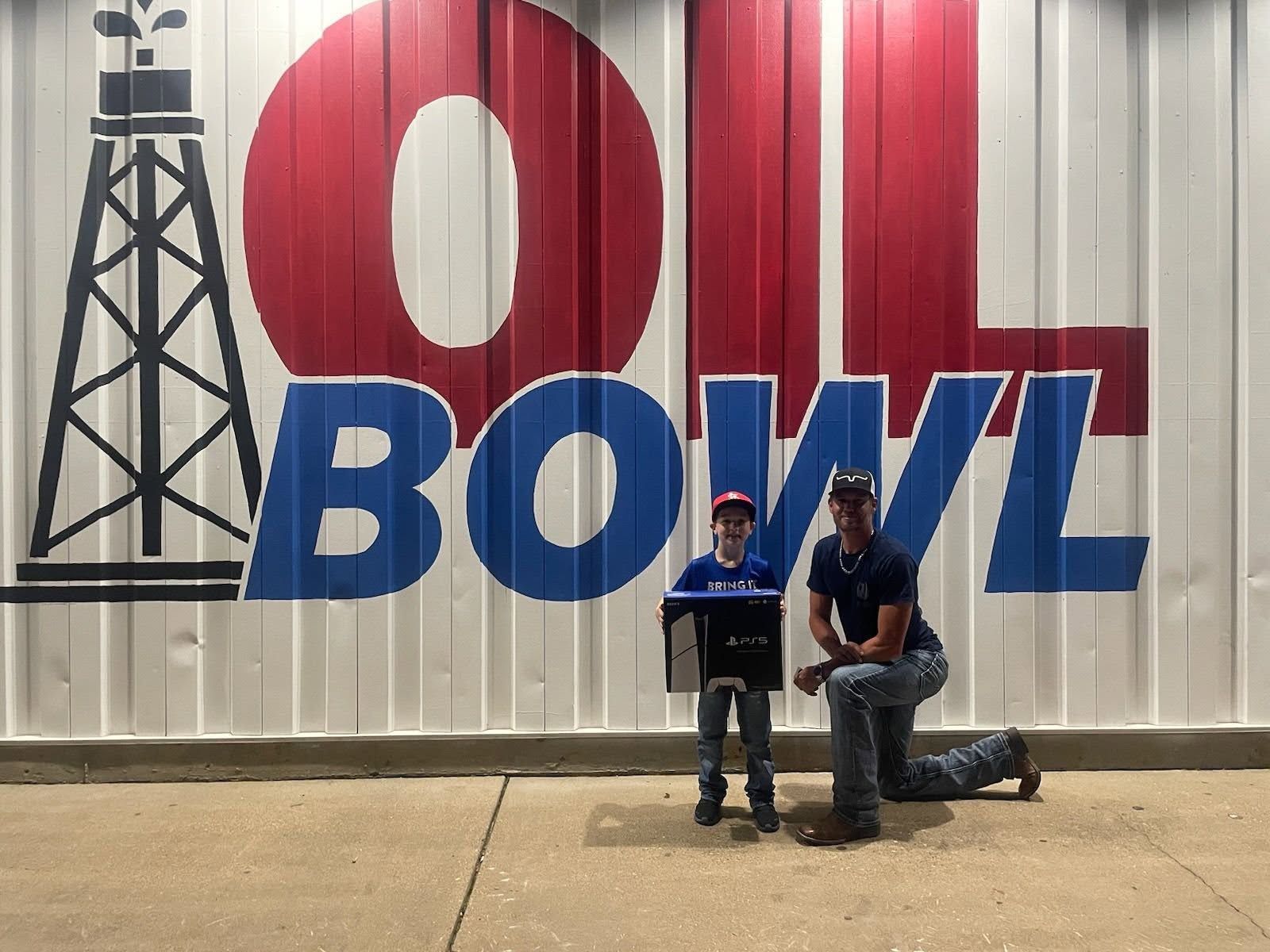 A boy holding a PlayStation 5 box and a man posing in front of a large red and blue Oil Bowl mural.